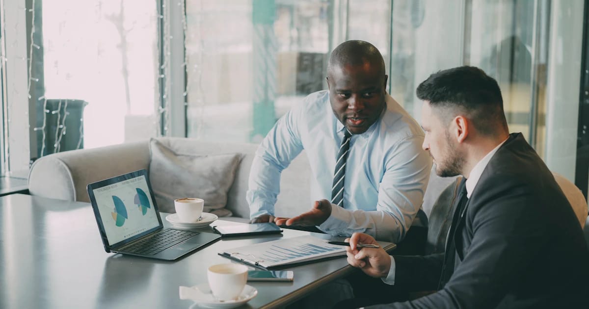 Hai người trao đổi về biểu đồ trên máy tính xách tay và tài liệu in tại bàn họp.